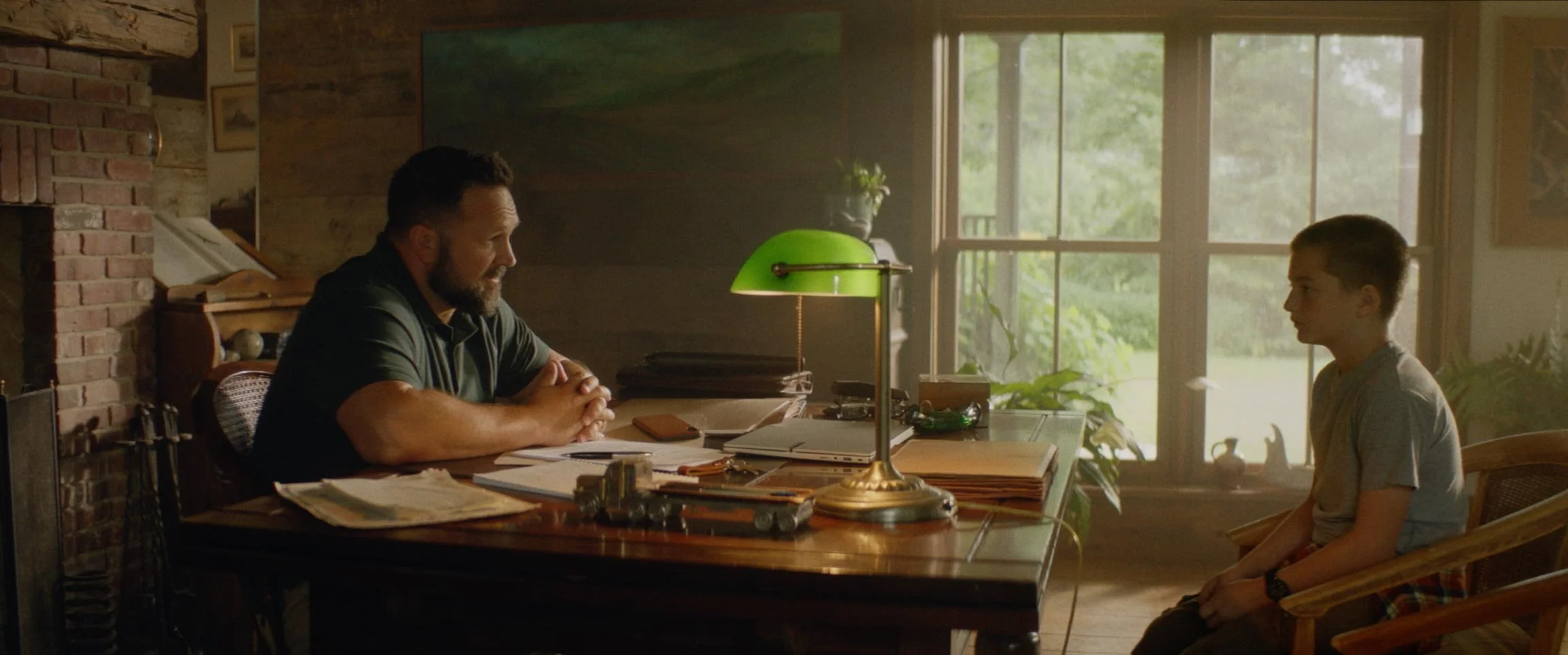 A man and boy sitting across from each other at a wooden desk in a cozy, rustic room with large window, green lamp, and plants, engaging in conversation.