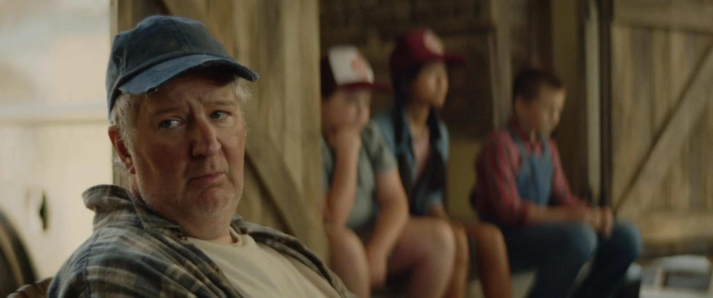 An older man wearing a plaid shirt and a cap sitting on a bench in a barn, with three young people in caps and casual clothes sitting behind him, all blurred.