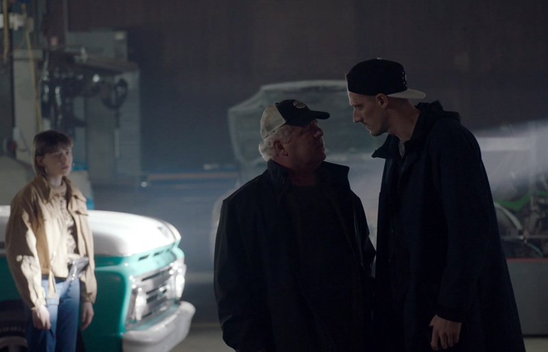 Three people standing indoors: a woman observing in the background, two men close together in the foreground engaged in a serious conversation. The setting appears to be a garage or workshop with vehicles in the background.