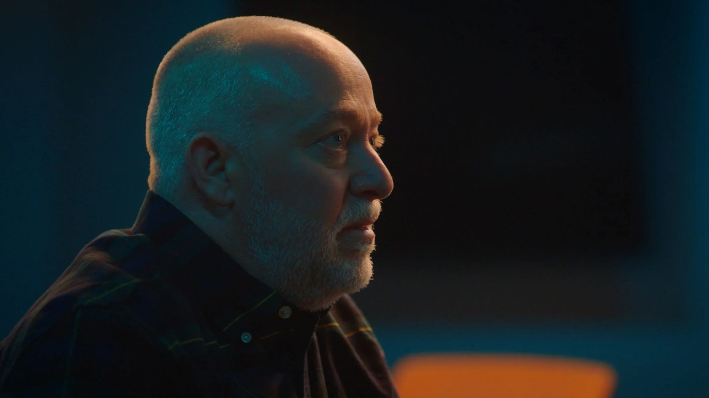 A profile of an older man with a bald head and gray beard, looking to the right, with dramatic, colorful lighting.