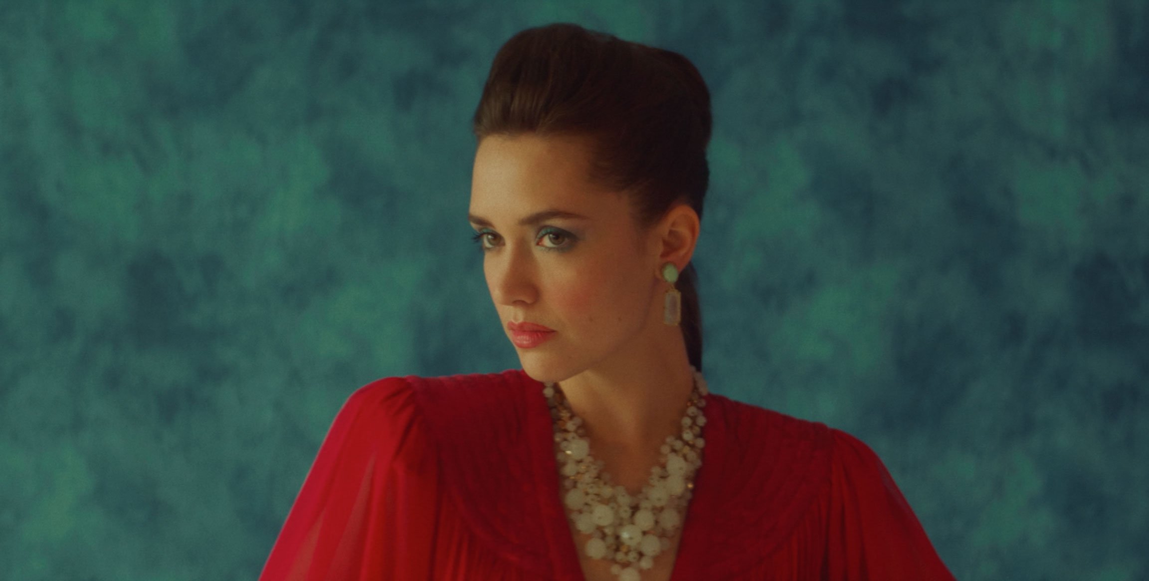 A woman with styled dark hair wearing a red dress and jewelry, looking over her shoulder against a dark green textured background.
