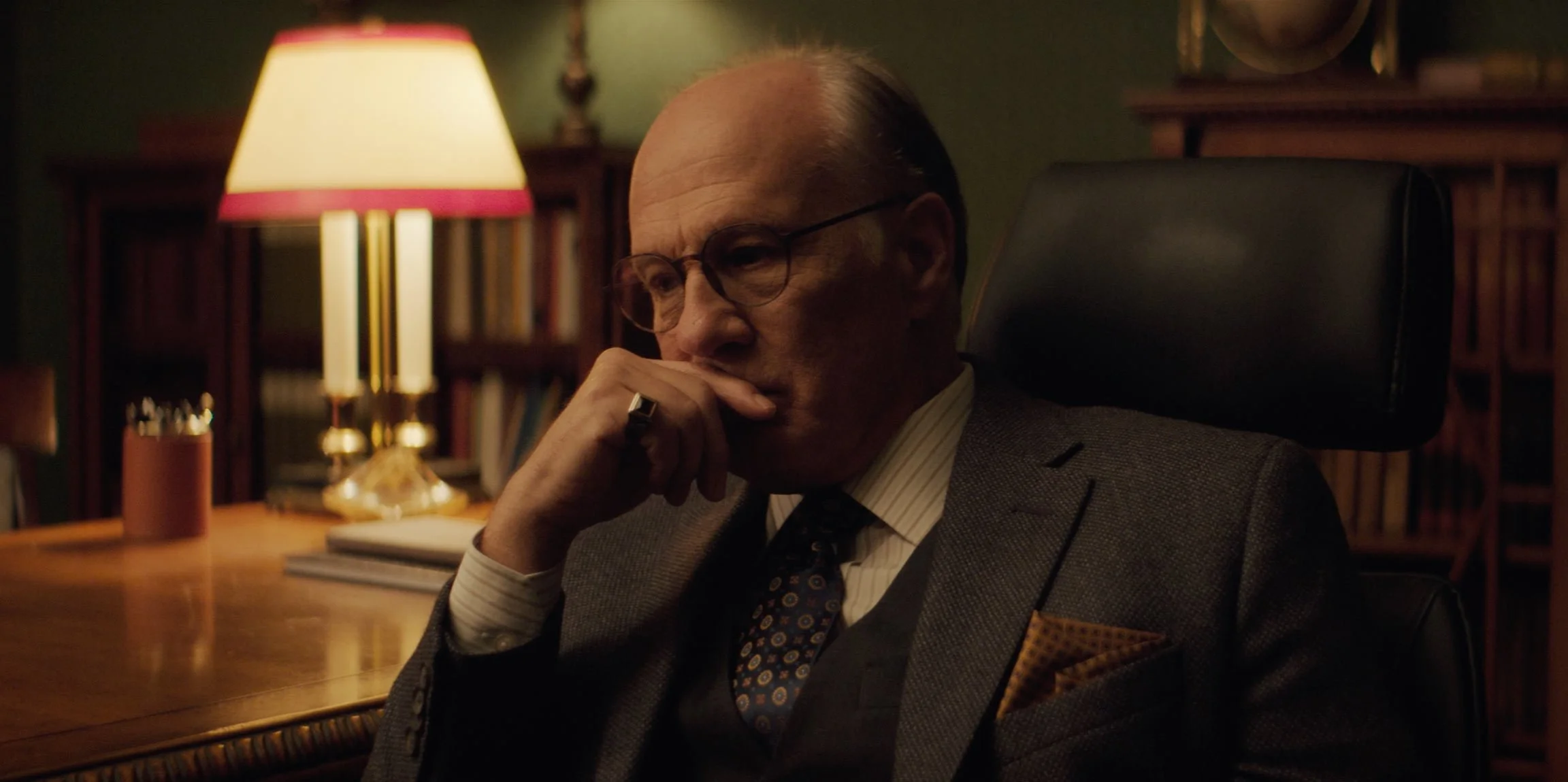 A man with glasses sitting at a desk in a bookshelf-lined room, deep in thought, with a lamp casting warm light behind him.