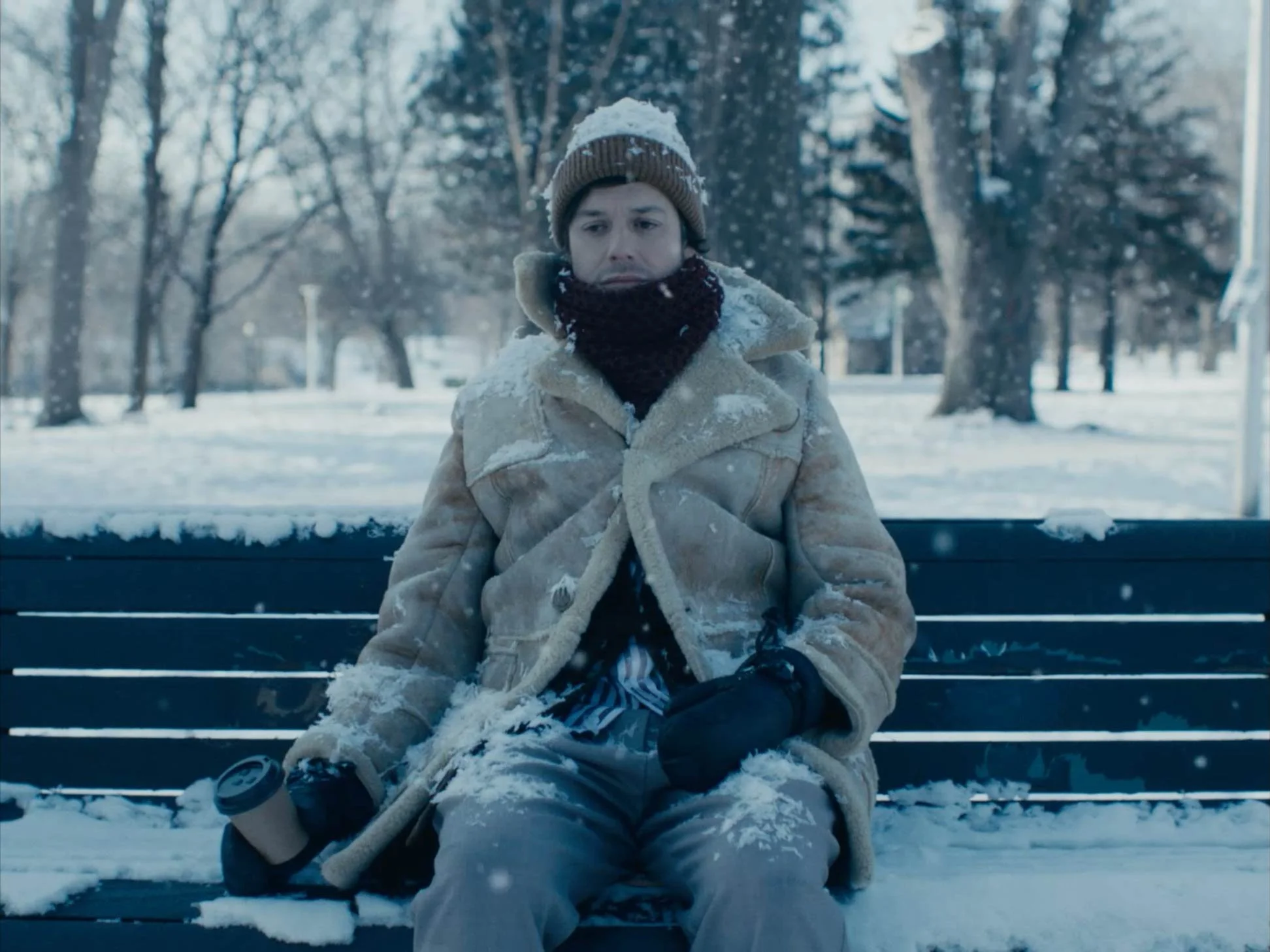 A man sitting on a snow-covered park bench in winter, wearing a beige coat, wool hat, black scarf, gloves, holding a coffee cup, with snow on his coat.
