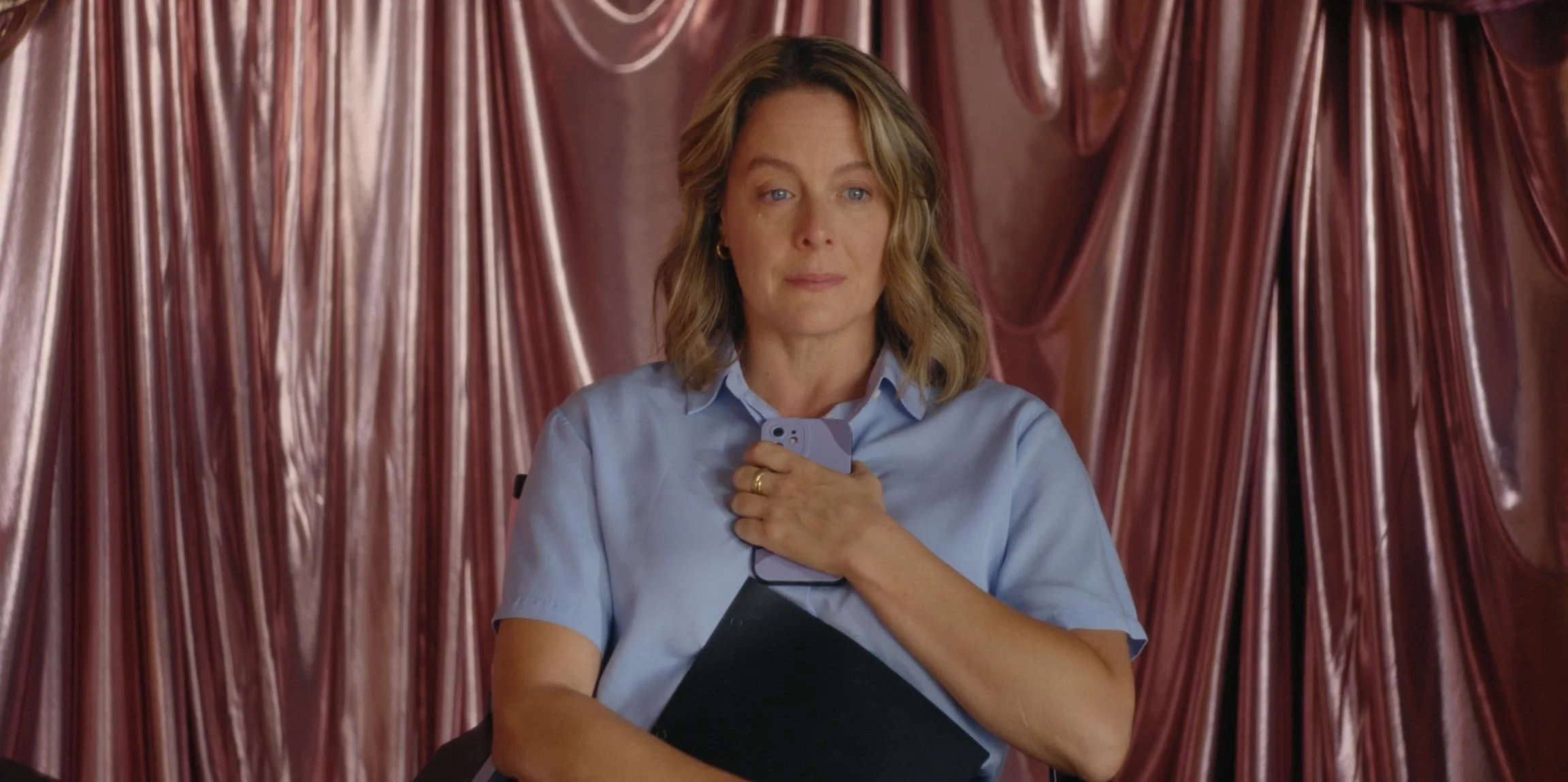 A woman with shoulder-length wavy blonde hair wearing a light blue shirt holding a smartphone with one hand against her chest, standing in front of a shiny pink satin curtain.