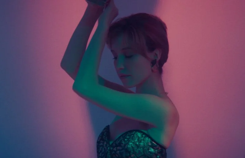 A young woman with short hair and closed eyes, posing with her arms raised above her head, illuminated by colorful neon pink and blue lighting.