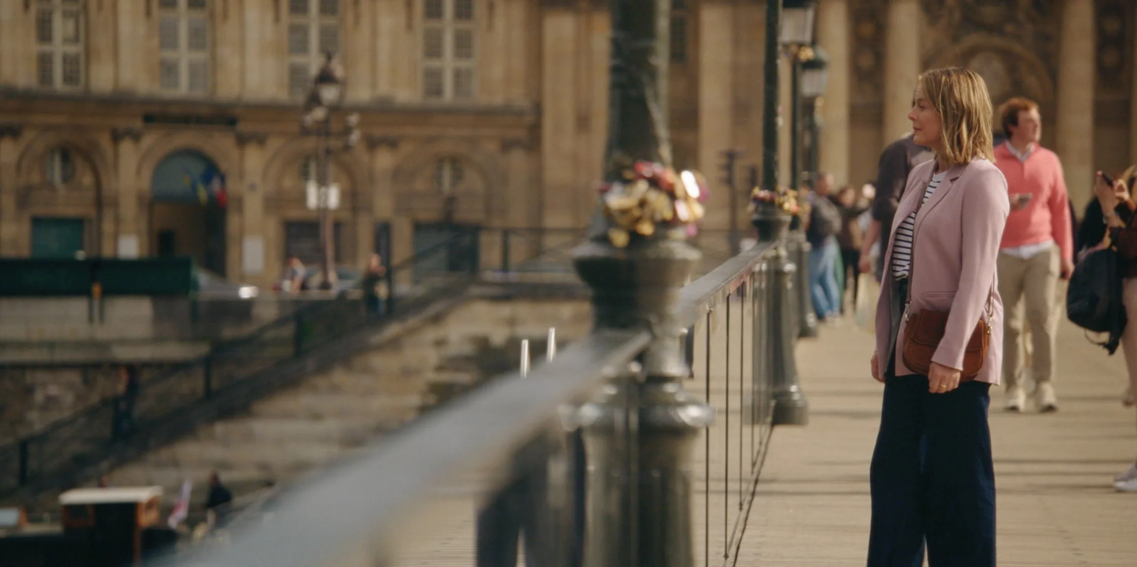 A woman with shoulder-length blonde hair wearing a pink blazer and striped shirt stands on a city bridge, looking to the right, with a brown bag over her shoulder. Other pedestrians walk past in the background, and historic buildings are visible.