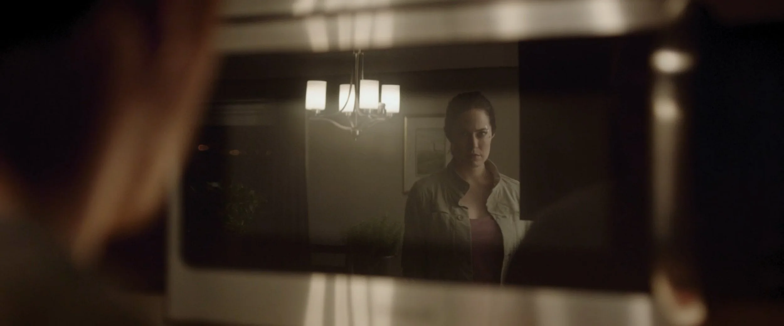 A woman looking at her reflection in a window or mirror, with a chandelier hanging above, in a dimly lit interior space.