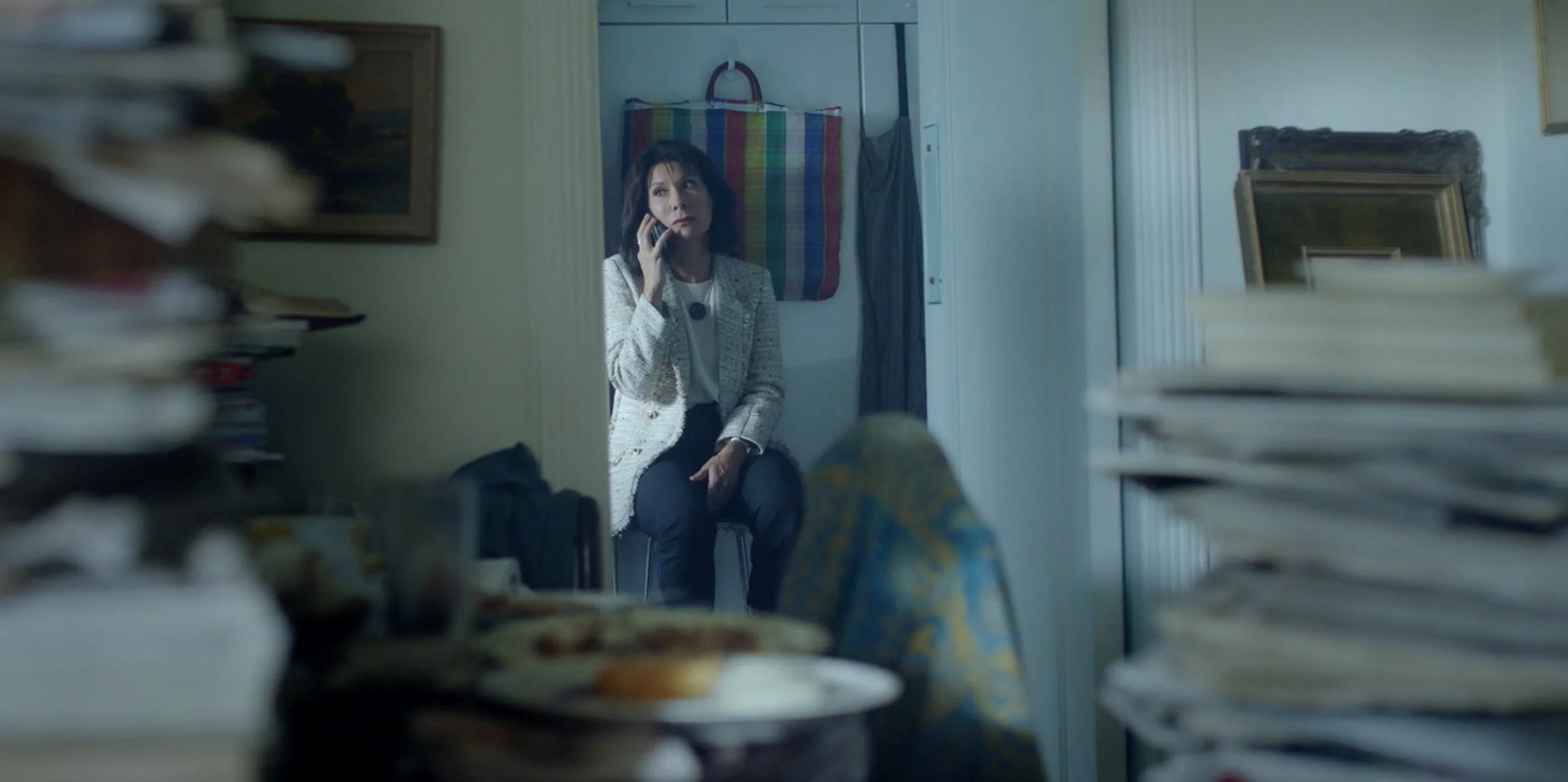 A woman sitting on a chair, talking on a cell phone, surrounded by stacks of papers and files in a cluttered room.