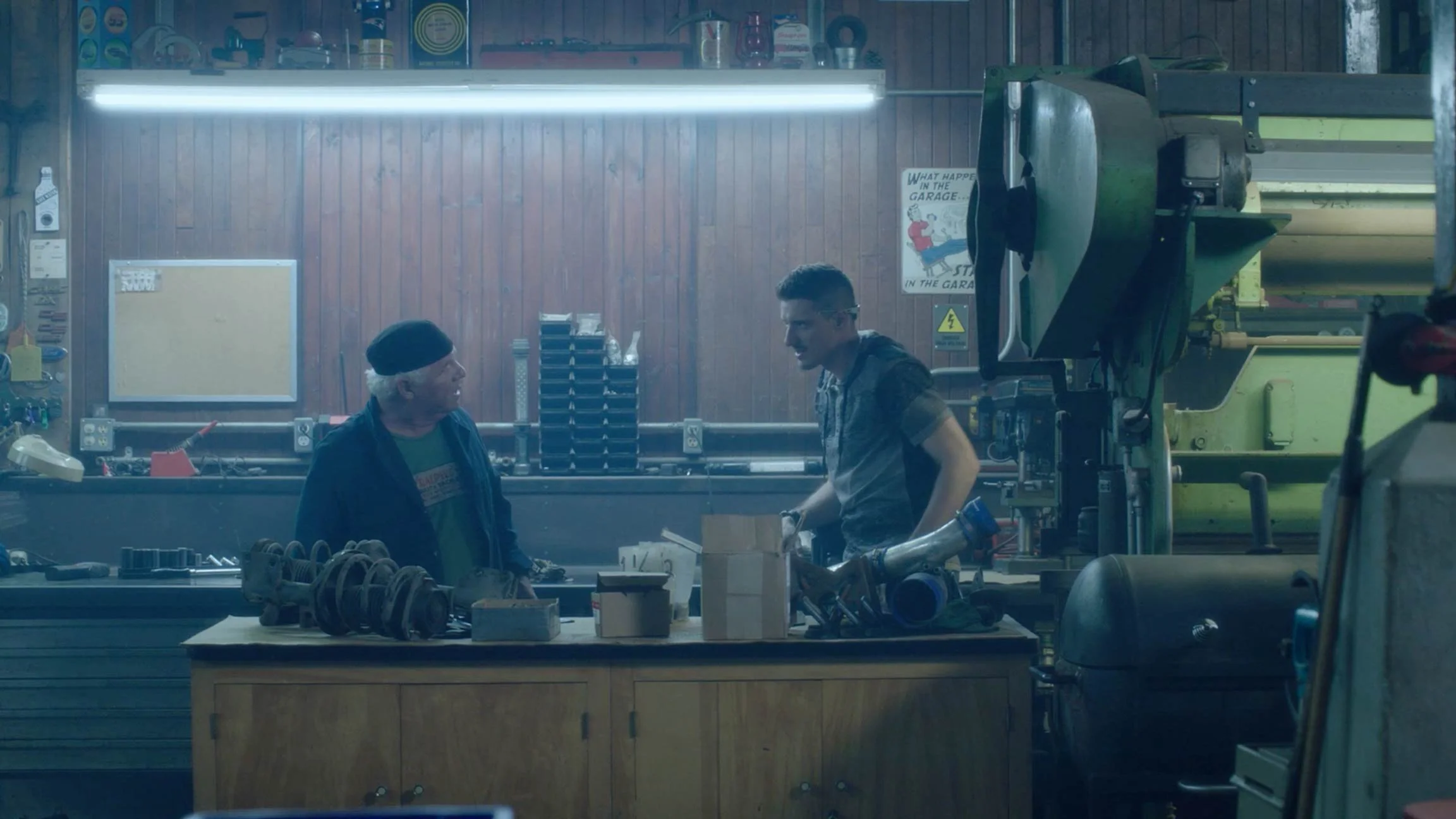 Two people in a garage workshop talking; an older man with white hair and a black beanie, and a younger man with dark hair and glasses, surrounded by tools and machinery.