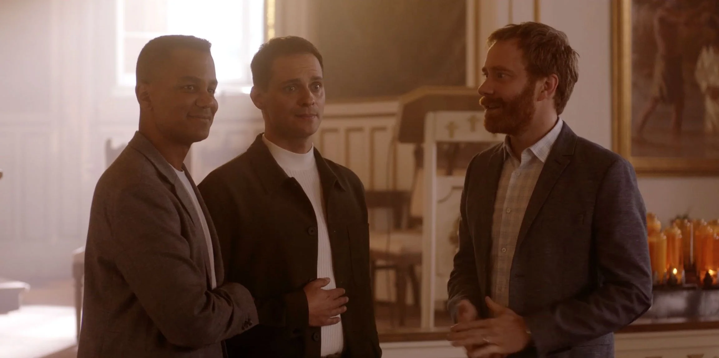 Three men standing and talking in a warmly lit room with paintings on the wall.