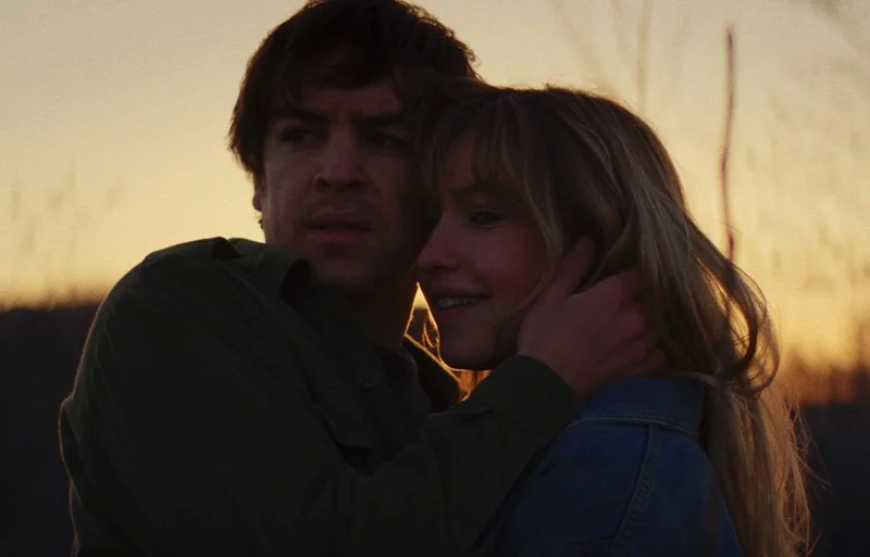 A man holding a woman close during sunset, both looking concerned or thoughtful.