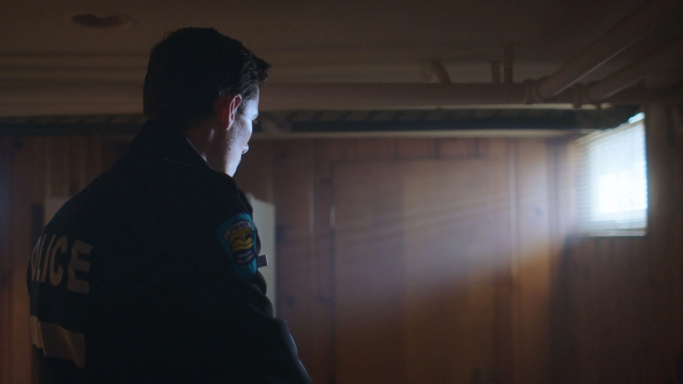 Side profile of a police officer in uniform inside a wooden-paneled room with a window, with sunlight filtering in.