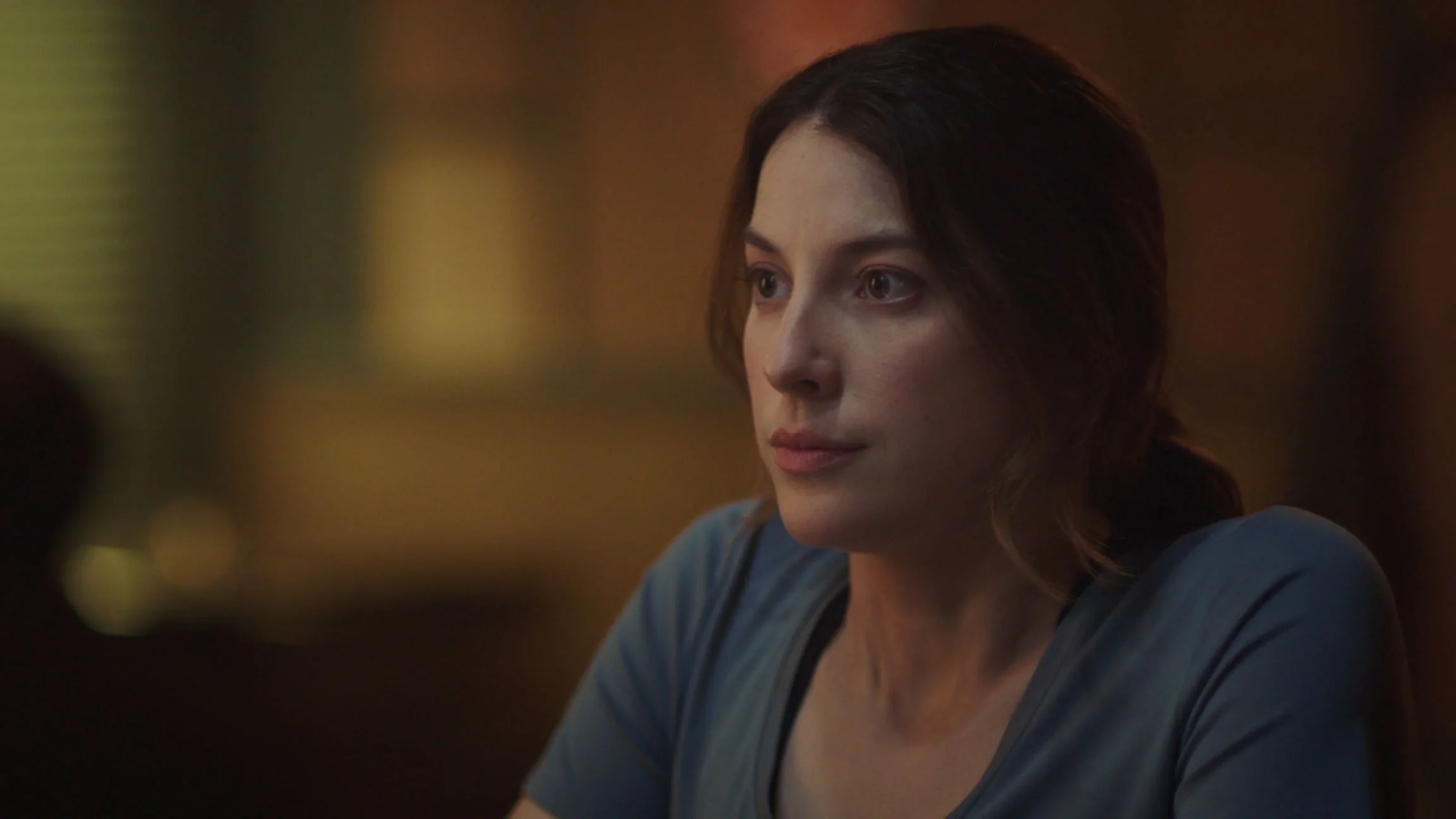 A woman with shoulder-length brown hair and a blue shirt looks to the side, with a thoughtful expression, in a dimly lit setting.