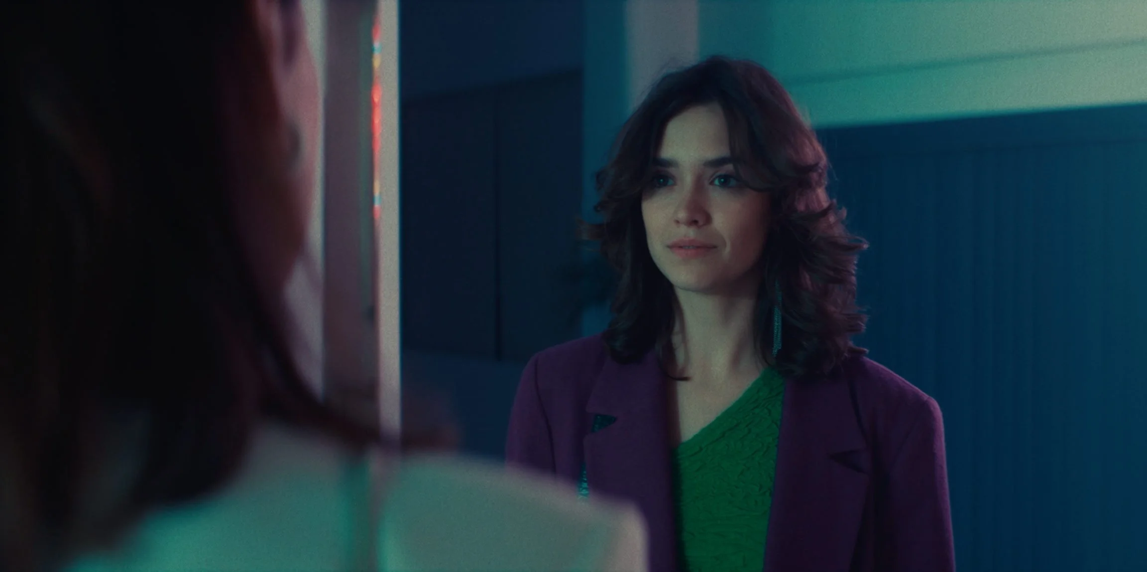 A woman with shoulder-length dark hair and makeup, wearing a purple blazer over a green top, looking at her reflection in a mirror.