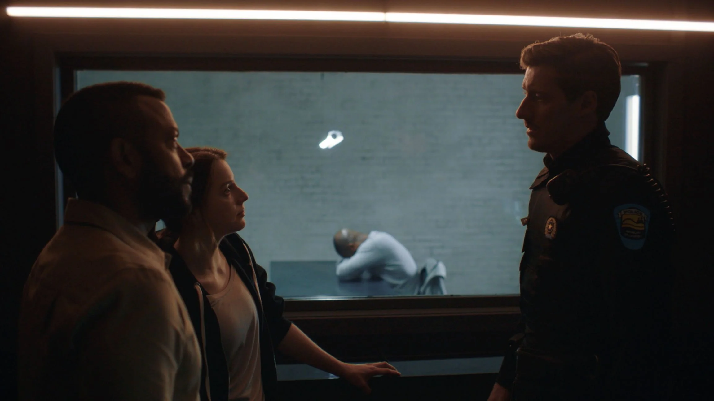 Three people, including a woman and two men, stand facing each other in a dimly lit room, with a police officer visible in uniform on the right side. A person in white is behind a glass window, sitting with their head bowed and their back curved, in 