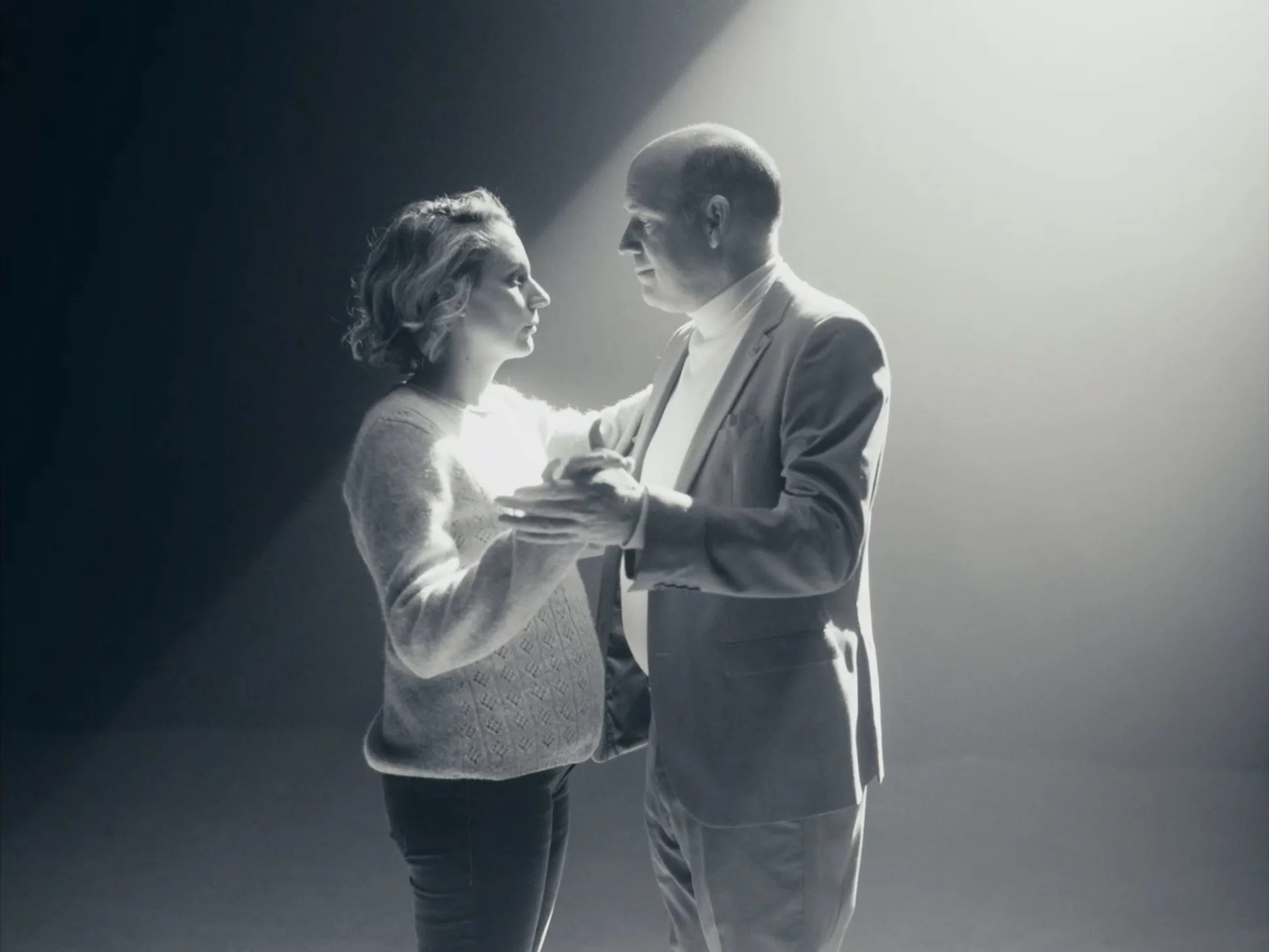 A black-and-white image of a man and woman facing each other, with the man holding the woman's hands, under a spotlight.