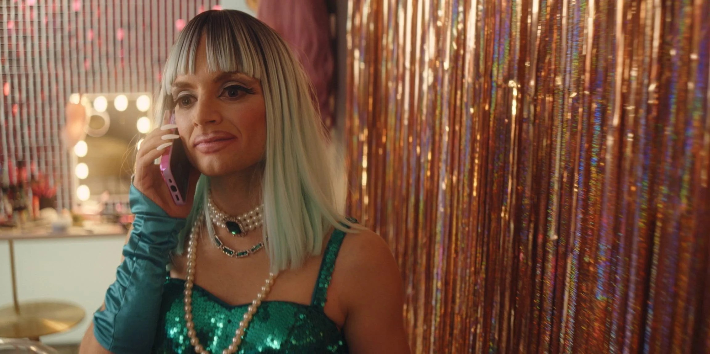 A woman in a sparkly green dress and layered pearl necklaces talking on a pink phone, standing in front of a glittering orange curtain.