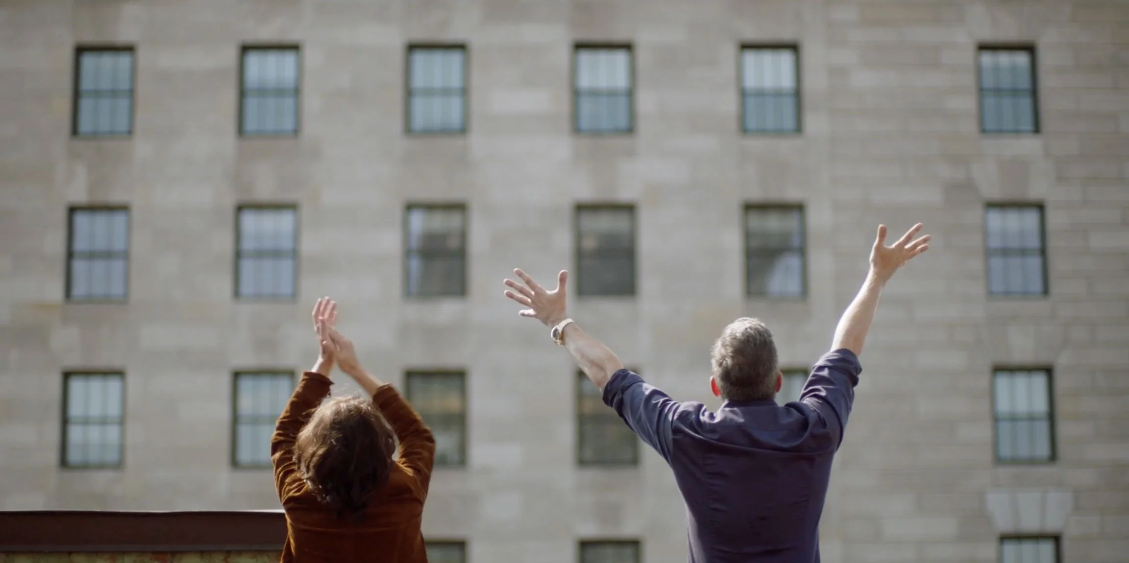 A man and a woman facing away from the camera with arms raised in front of a multi-story building with multiple windows