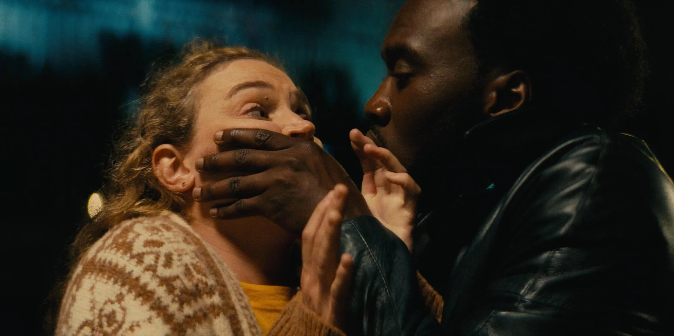 A Black man is holding a woman's face with his hand while she looks surprised or scared. The scene appears tense and emotional, set against a dark background.