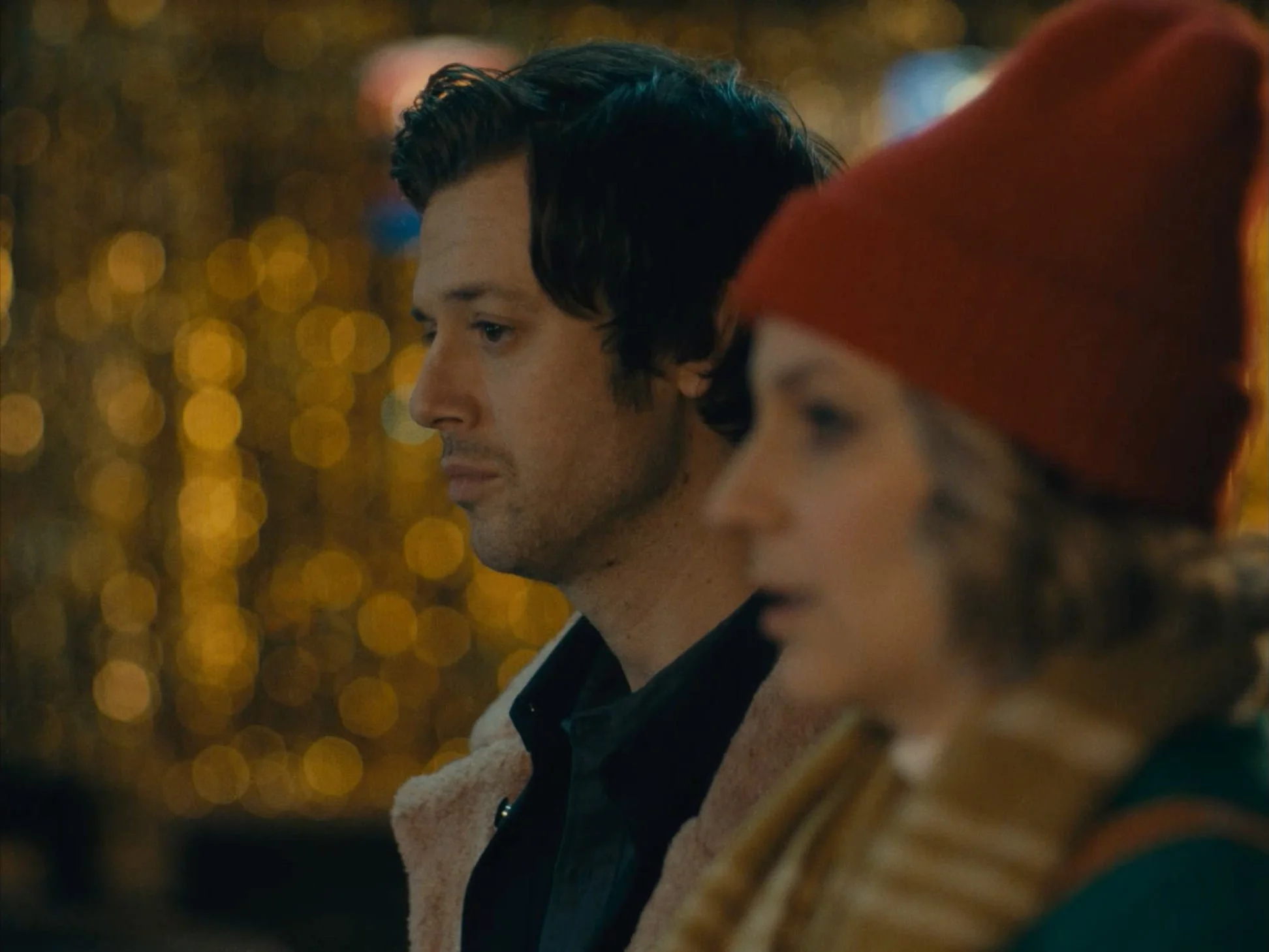 A man and a woman in winter clothing standing near each other at night, with blurry golden lights in the background.