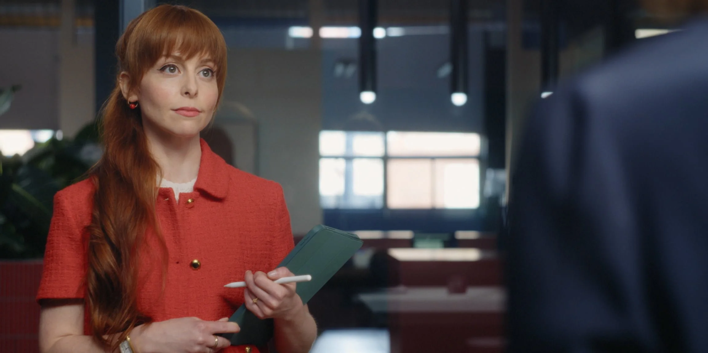 A woman with long red hair, wearing a red coat and holding a notepad and pen, standing indoors and engaging in conversation.