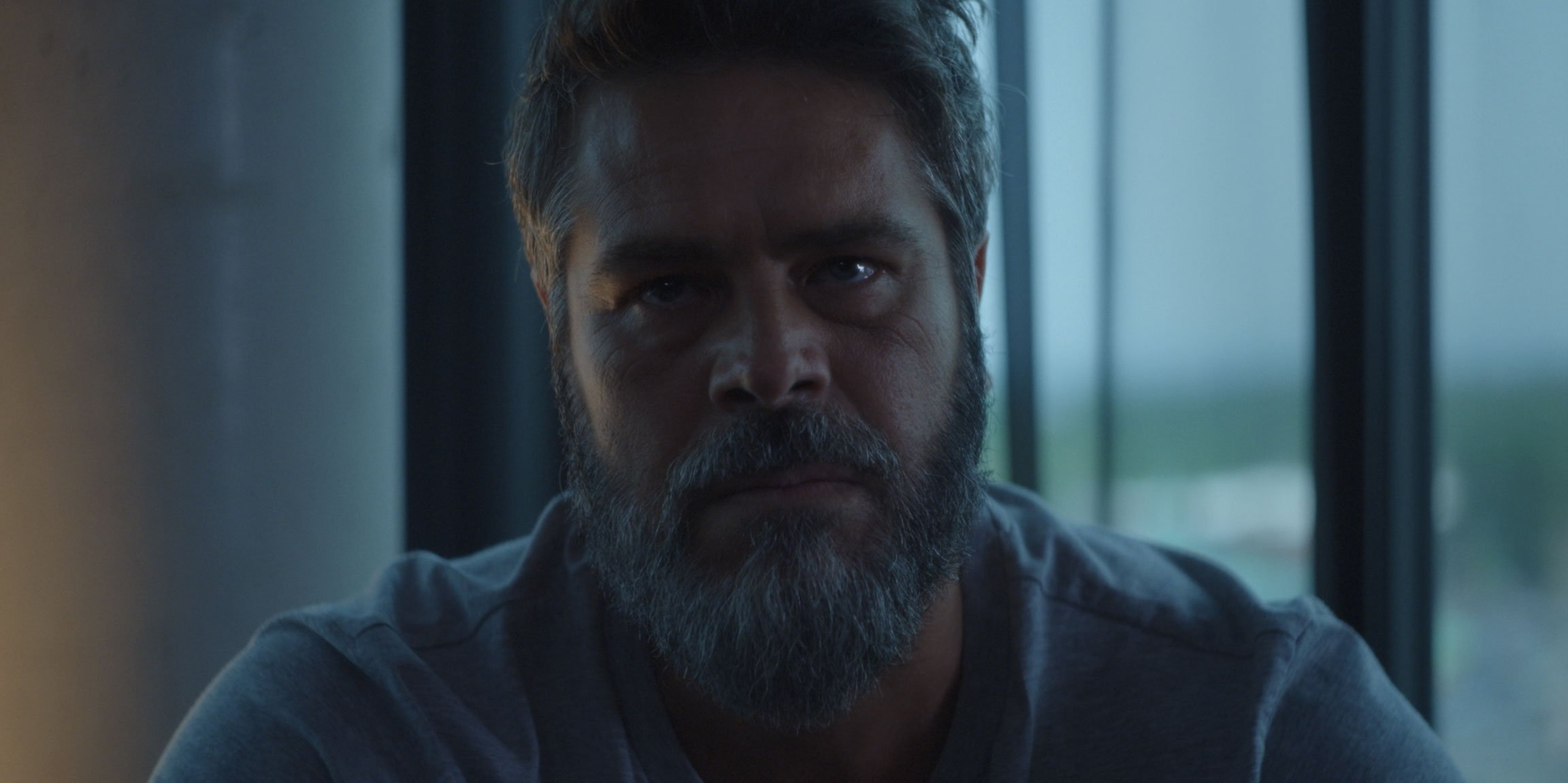 Close-up of a man with a beard and serious expression sitting indoors near a window.