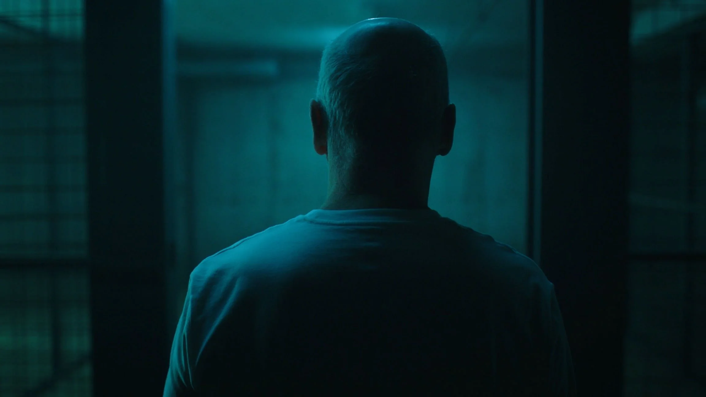 Back of a person with a shaved head, facing away, in a dark, teal-lit space that resembles a hallway or corridor.