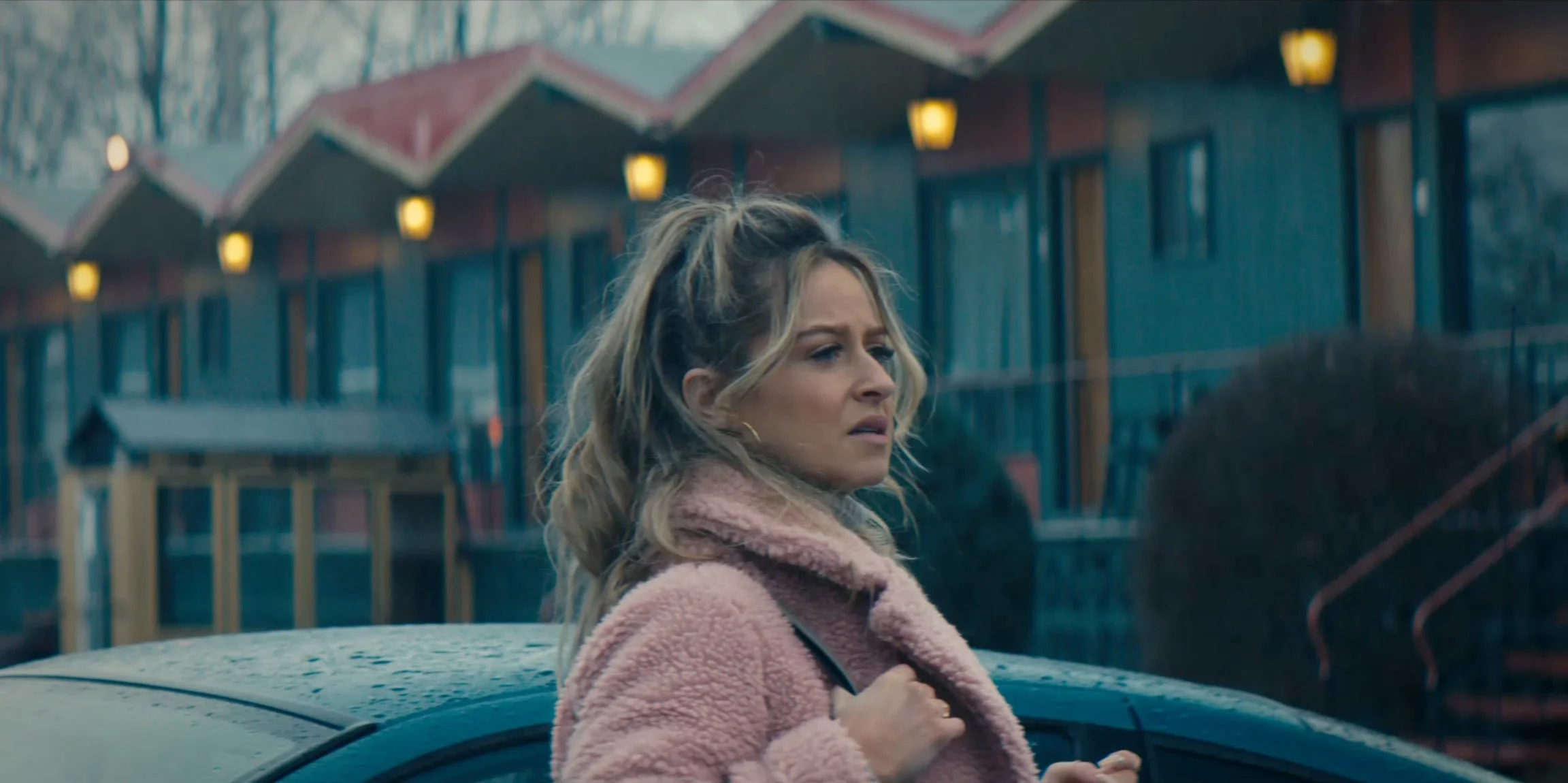 A woman with blonde hair in a pink fluffy coat standing outside on a rainy day next to a car, with a building with warm lights in the background.
