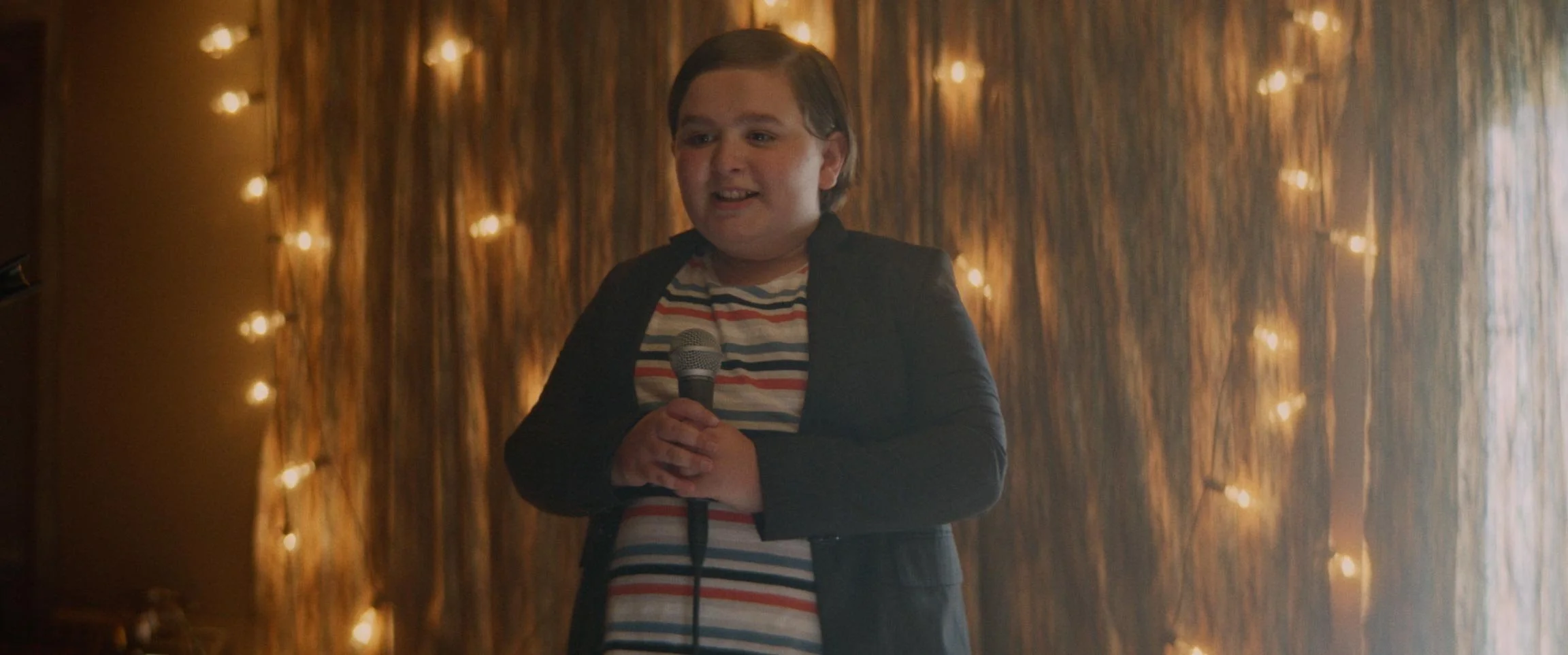 A young boy holding a microphone, standing in front of a wooden background decorated with string lights, smiling.