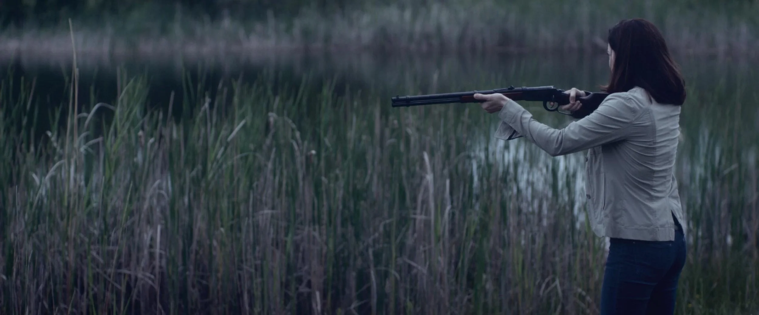 A woman aiming a rifle in a marshy area near water with tall grass.
