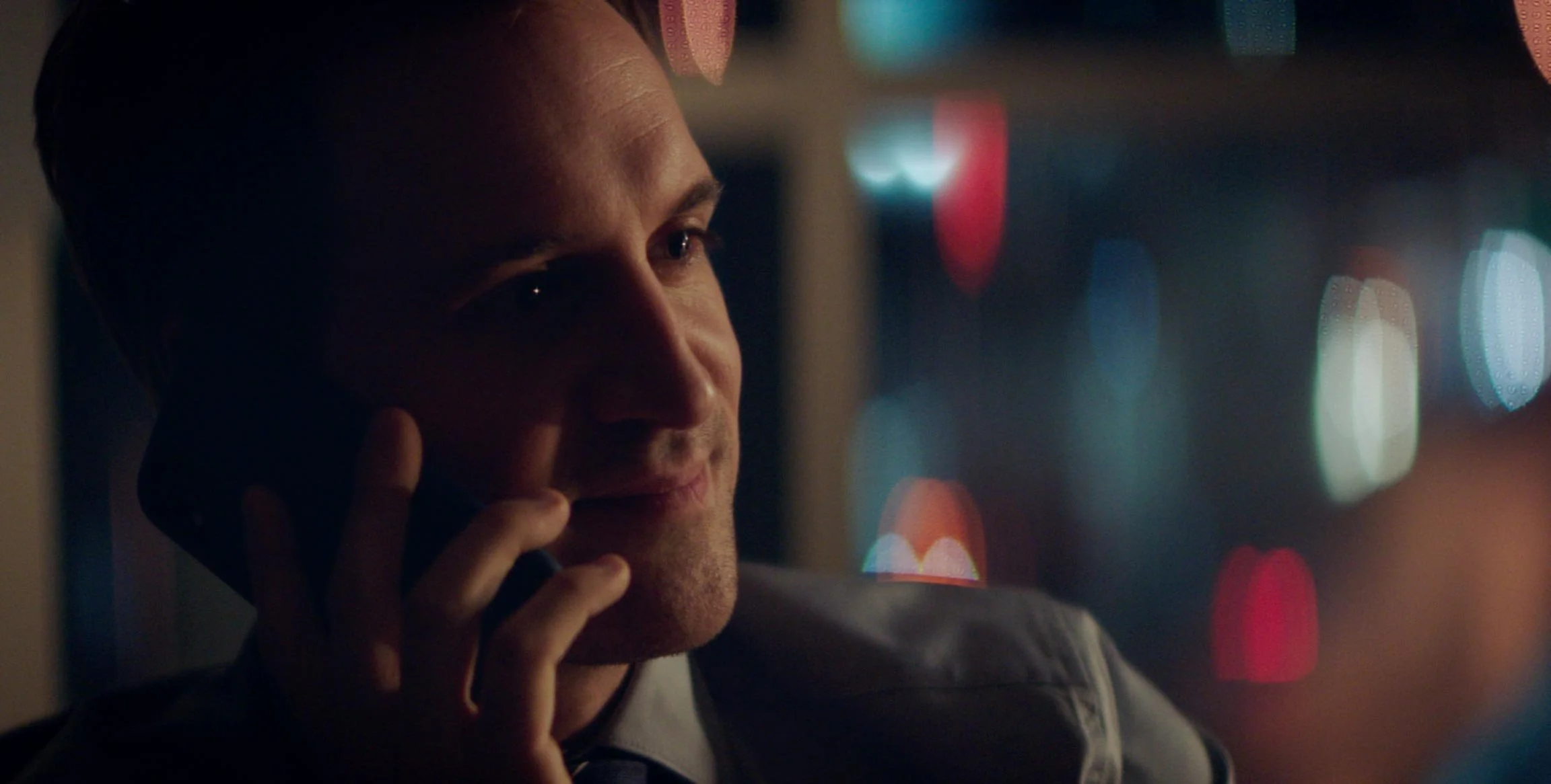 A man talking on a cellphone at night, with colorful city lights out of focus in the background.