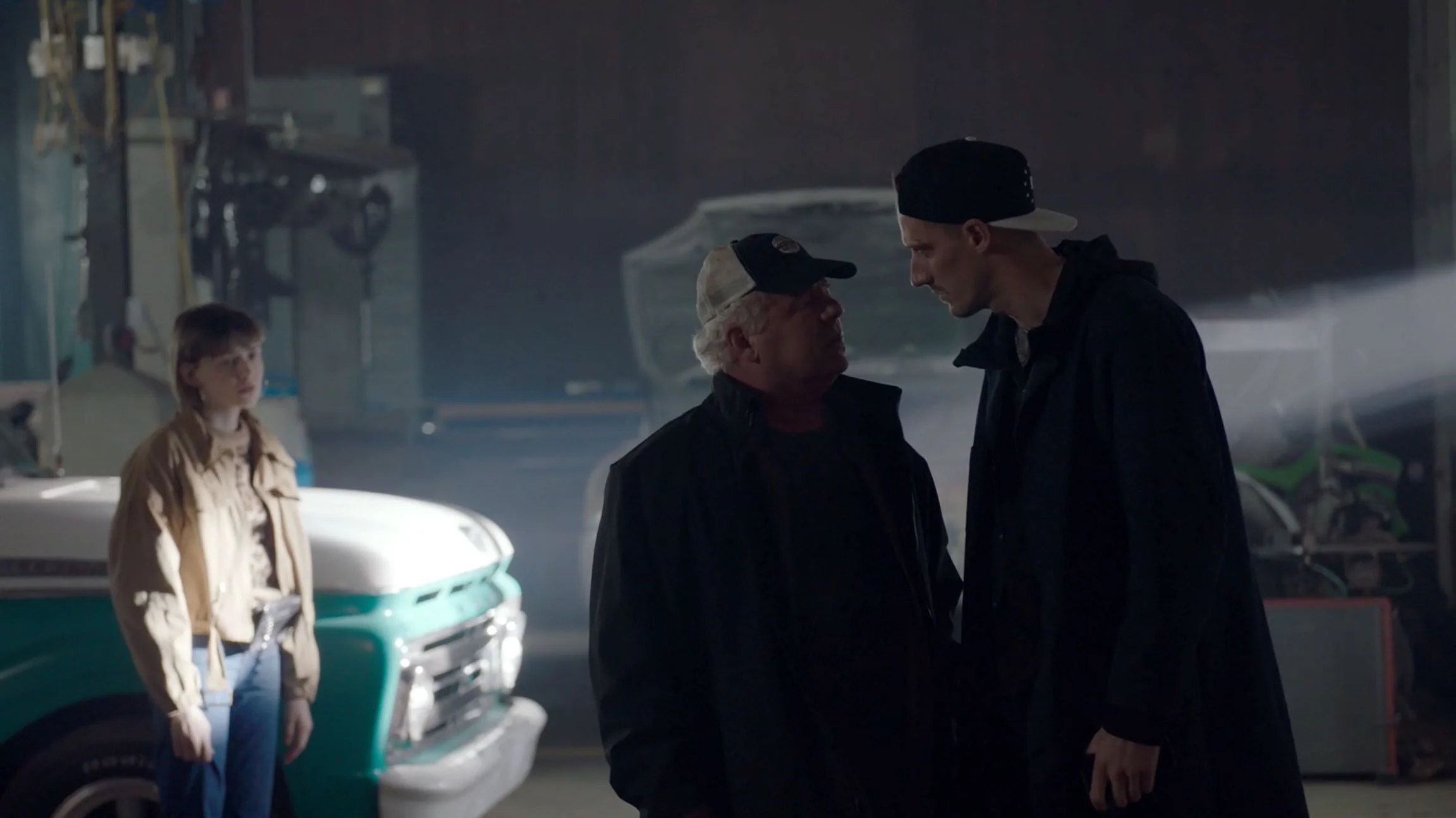 Three people in a garage: a young woman standing near a vintage green and white truck, and two men facing each other, engaged in conversation.