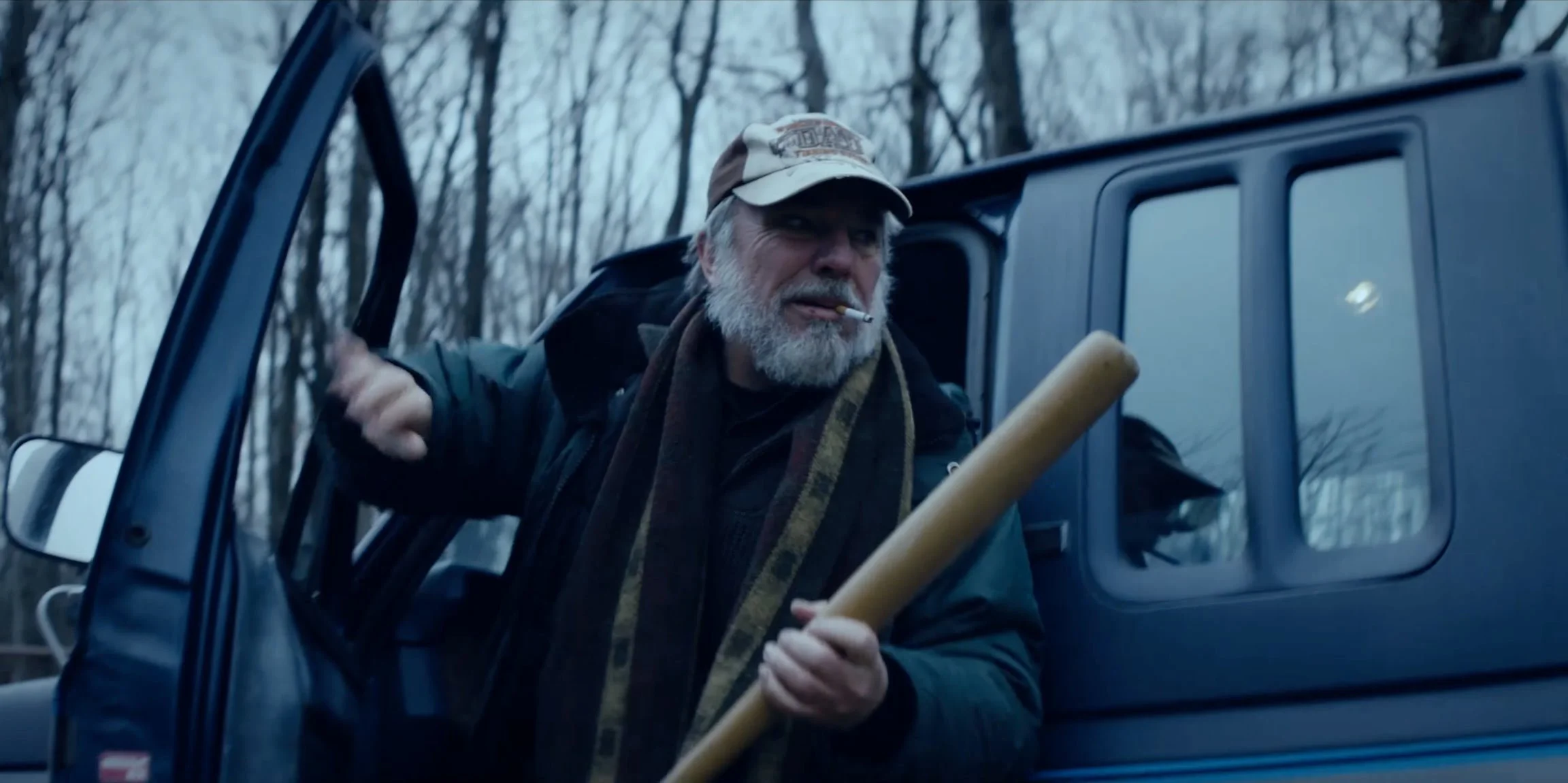 An older man with a white beard, wearing a beige cap, smoking a cigarette, holding a baseball bat, and getting out of a black vehicle in a wooded area.