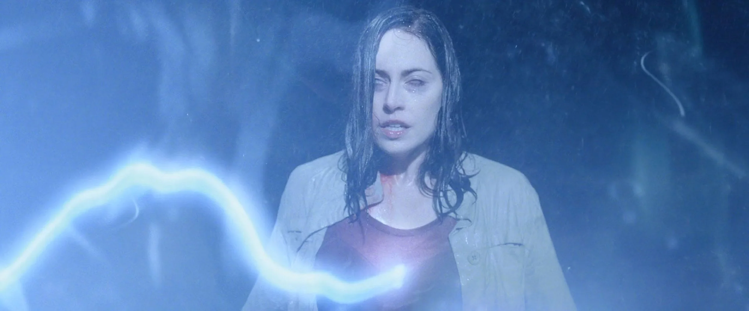 A woman with wet hair, wearing a beige jacket over a red shirt, is standing behind a blue lightning bolt, with a somber expression.