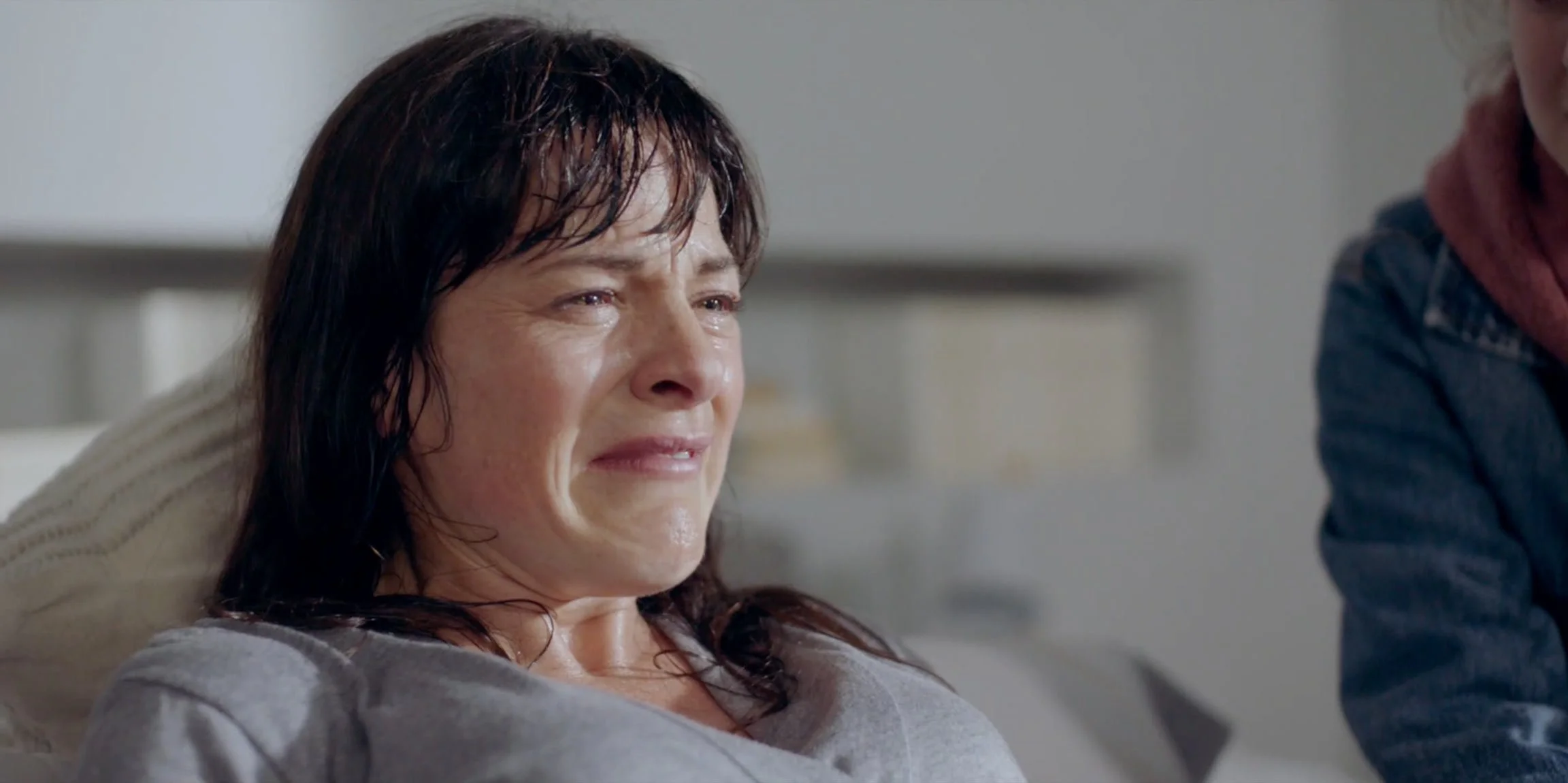 A woman with wet, dark hair crying and visibly upset while sitting inside a room.
