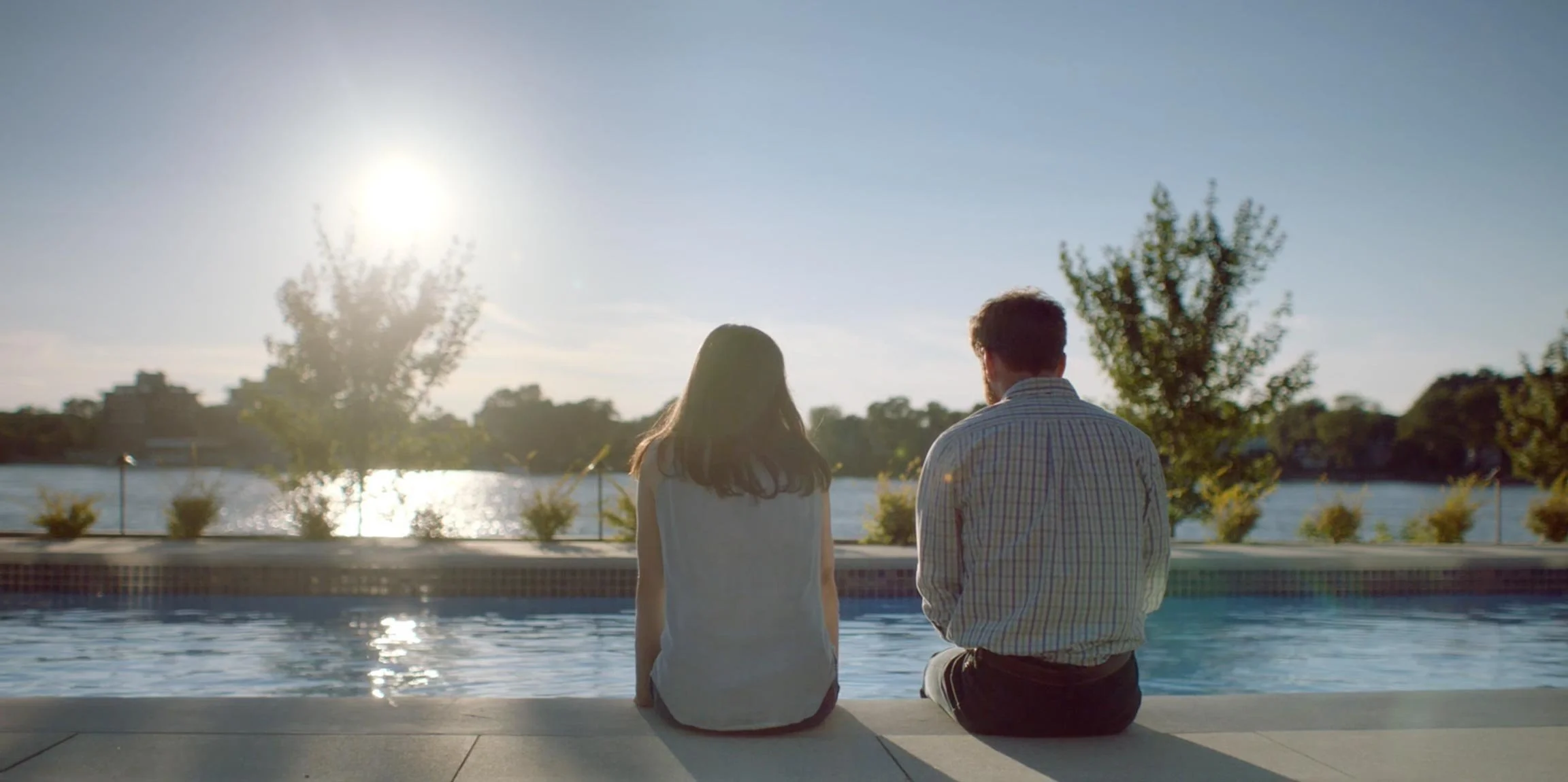 A man and woman sitting by a pool, facing a river with trees and buildings in the background, during sunset or sunrise.