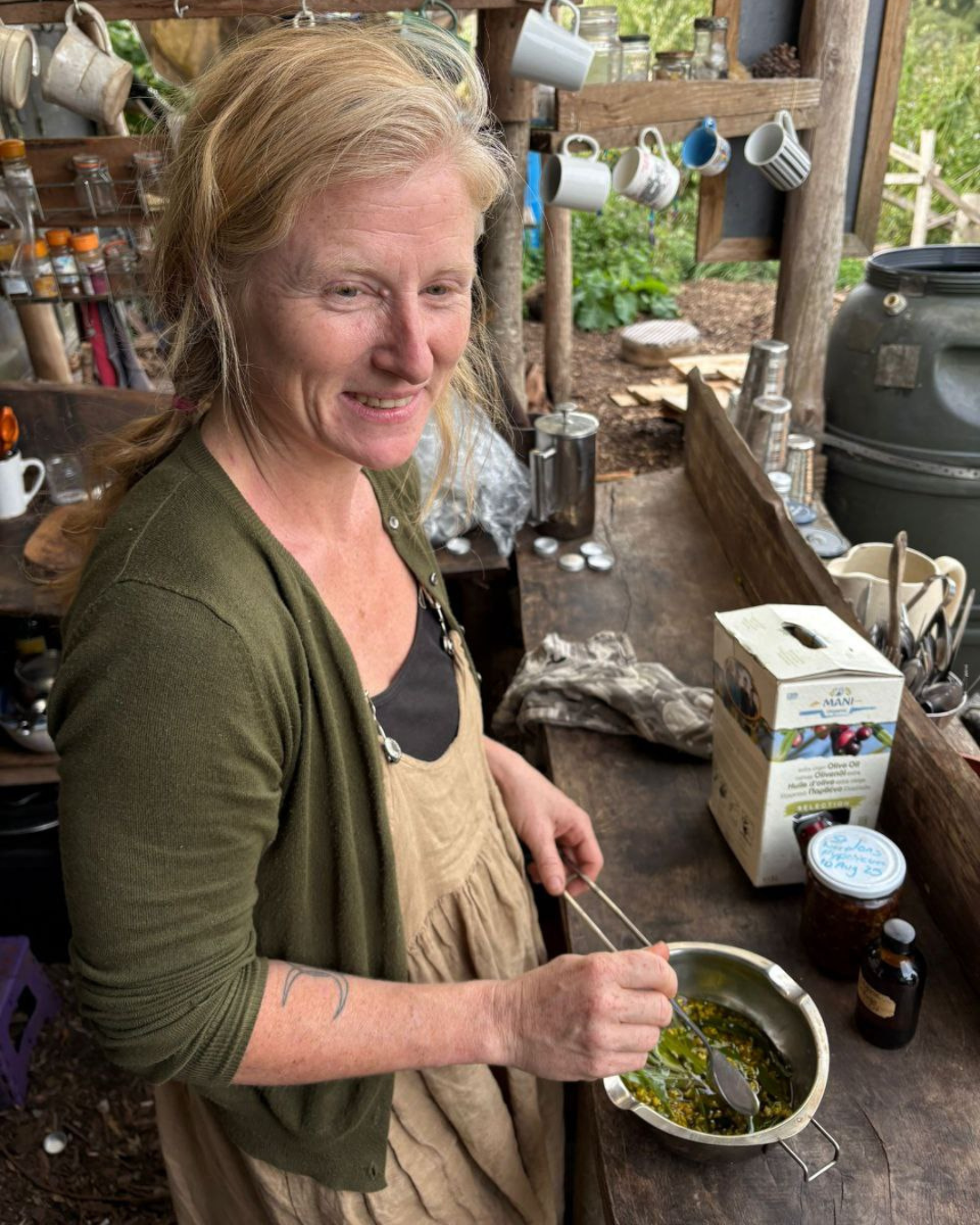 A woman with blonde hair and a tattoo on her left arm preparing herbal oil infusion in an outdoor rustic kitchen.