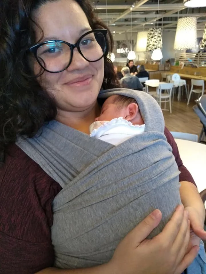 Woman holding a newborn baby in a baby carrier at a indoor café, with a background of tables, chairs, and hanging lamps.
