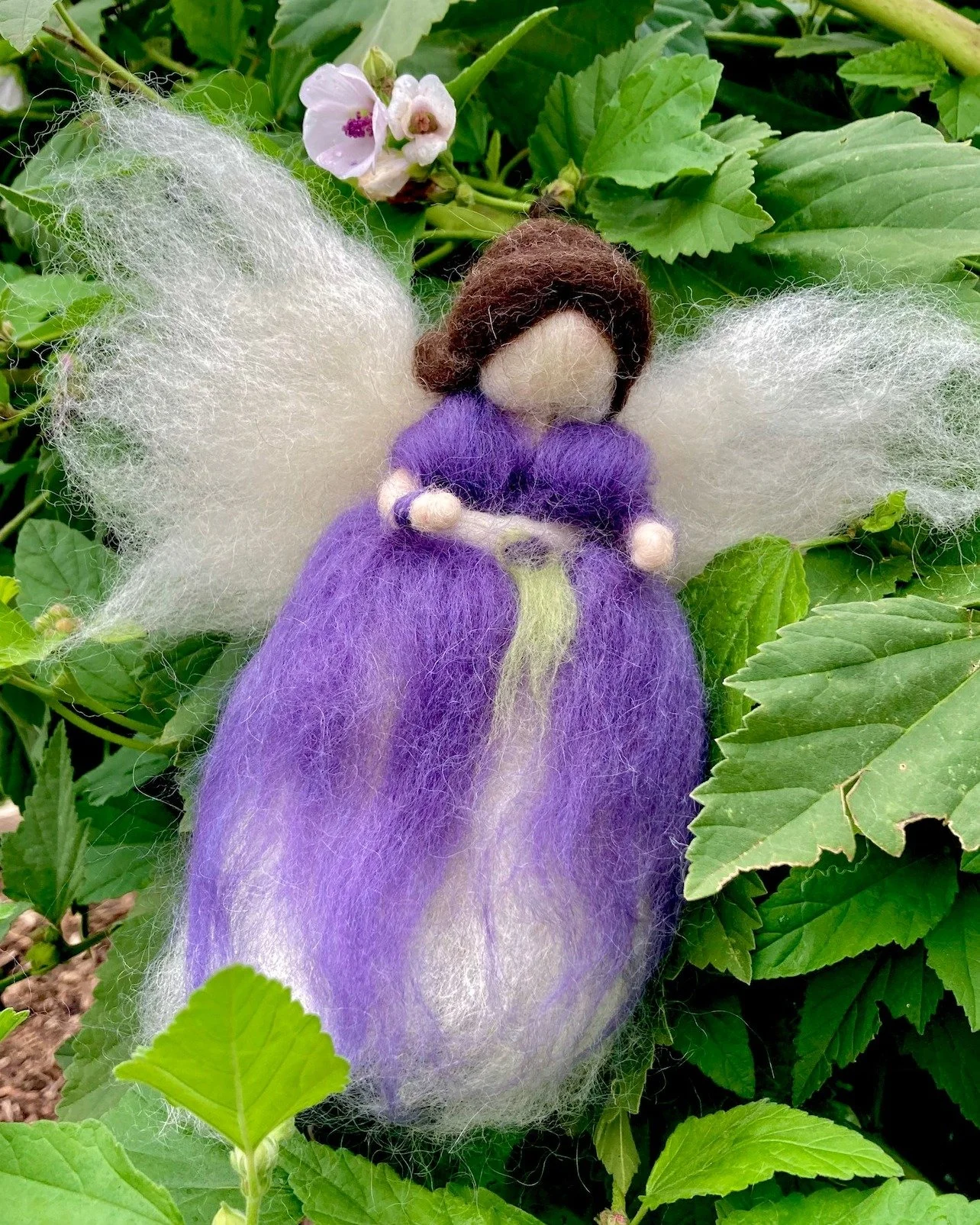 Felted fairy figurine with purple dress, white wings, and brown hair, sitting among green leaves and pink flowers.