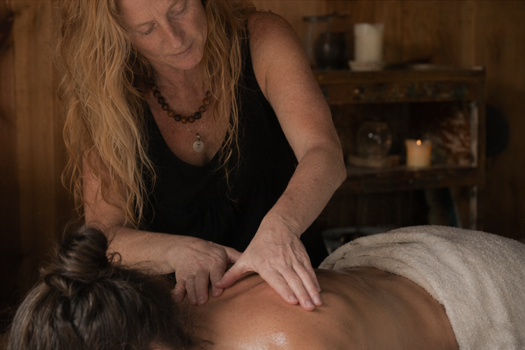 A woman giving a massage to a person lying face down on a massage table, in a cozy wooden room with natural light and plants.