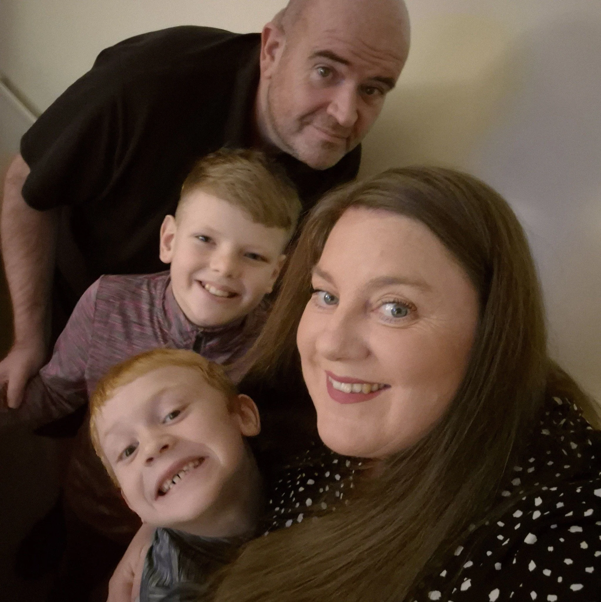 A family taking a selfie together indoors, including a woman with long brown hair, two young boys with red hair, a man with a shaved head, all smiling and close to each other.