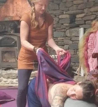 A woman in a brown dress and gray tights is performing a massage or therapy on a woman lying on a mat, using a purple blanket. They are indoors near a stone wall with a fireplace.