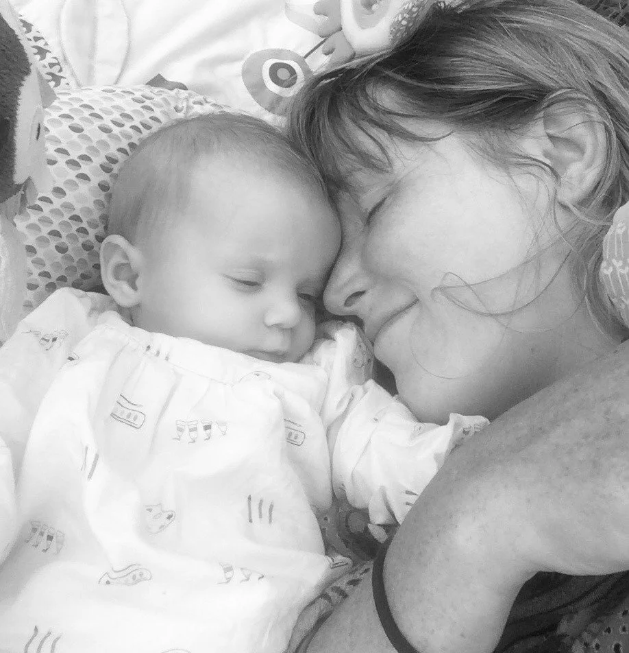 A woman with light-colored hair and a young child with a bald head are lying close together, touching foreheads and smiling with closed eyes.