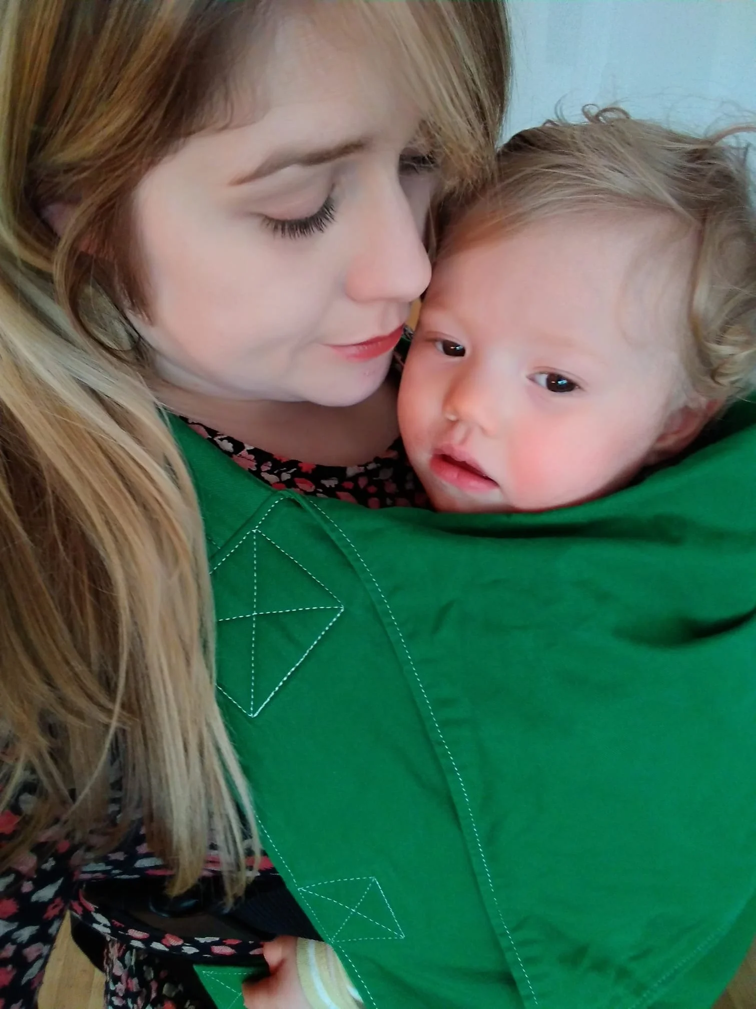 A woman with light skin and long blonde hair embracing a young child with curly blonde hair and light skin, both with closed eyes, sharing a tender moment.