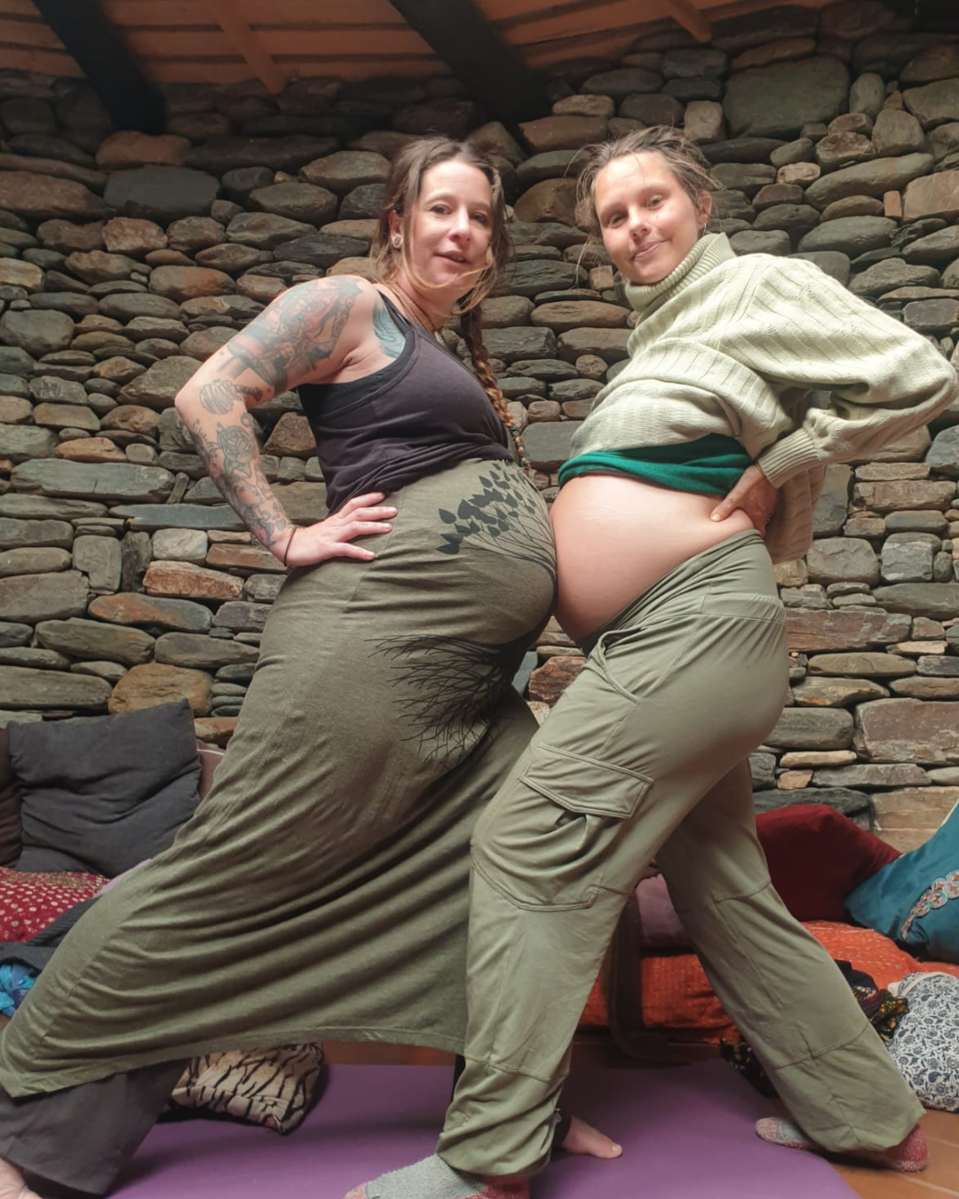 Two women, one pregnant with a large belly, pose together in front of a stone wall, smiling with hands on hips.
