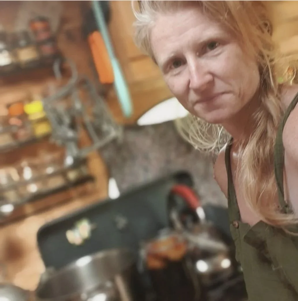 A woman with long blonde hair in a kitchen, near a stove, with wooden cabinets and shelves in the background.