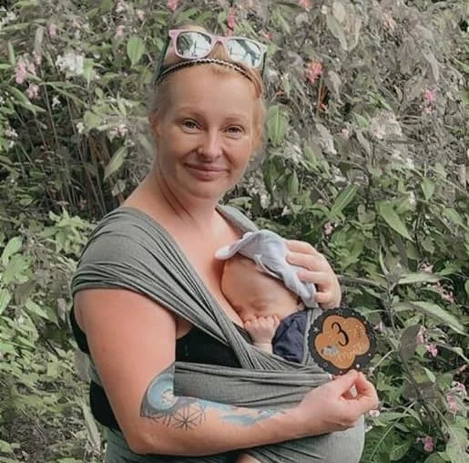 A woman with light-colored hair and sunglasses on her head is smiling and carrying a baby in a sling on her chest. The woman has tattoos on her arm, and the background is filled with greenery and pink flowers. The baby is resting with a hand on the woman's chest and is wearing a hat.