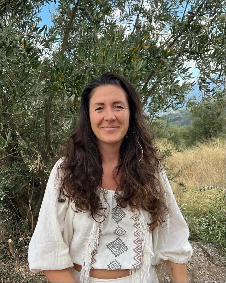 Woman with long dark wavy hair smiling outdoors, standing in front of green foliage and trees, wearing a white embroidered top with fringe details.