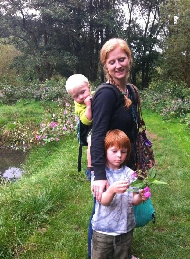A woman with two children outdoors on a grassy trail surrounded by trees and purple flowers. One child, with red hair, is inspecting a flower. The other child, a toddler, is on the woman's back, smiling and sucking on their fingers.