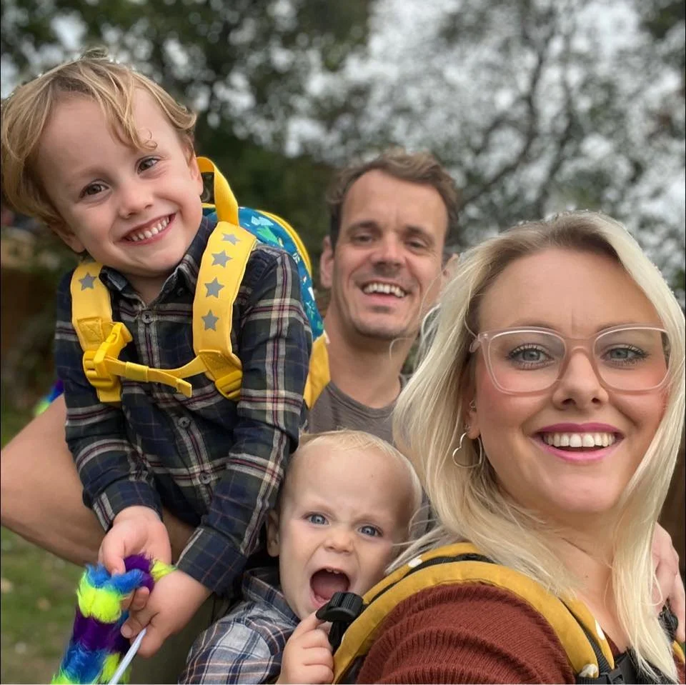 A family of four outdoors, smiling at the camera. The mother has blonde hair and wears glasses, while the father has brown hair. The boy on her back is smiling, and the younger child in front appears surprised or excited. They are all wearing casual clothing and carrying backpacks, with trees in the background.